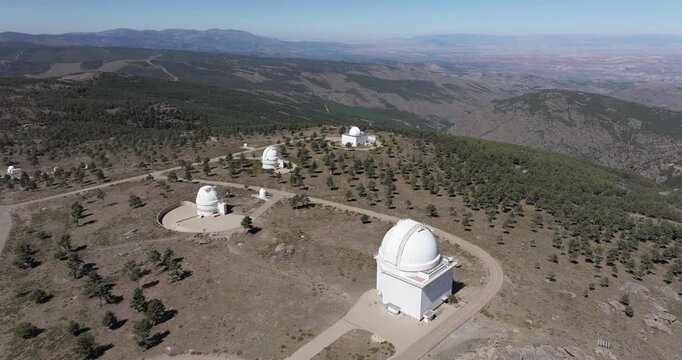Wide drone shot of Calar Alto Observatory with multiple telescope domes and winding access roads across rocky plateau in Spain