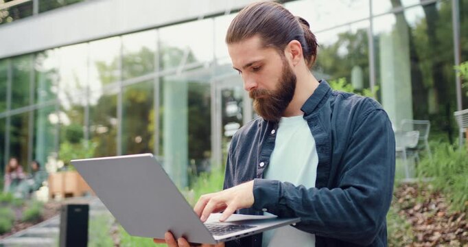 Handsome entrepreneur using a wireless laptop outdoors near a modern office building, typing and scrolling while managing online business and remote work.