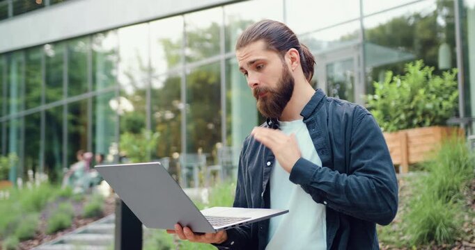 Successful bearded businessman greeting colleagues with hand gesture during online conference on laptop outdoors, working remotely. Concept of modern flexible work and global digital communication.