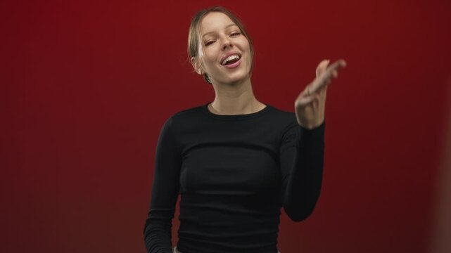 Woman smiling with open right hand reaching toward camera wearing black long sleeve top in red studio; playful charm.