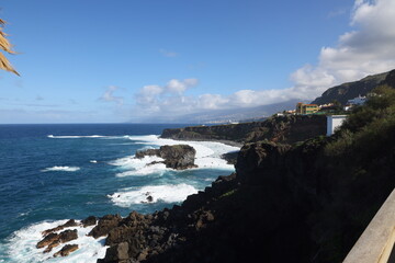 Obraz premium Dramatic coastal view from Tenerife’s northern cliffs with deep blue Atlantic, crashing waves and distant cliffside buildings under sunlit sky
