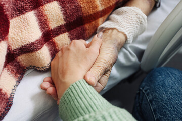 Young woman holding elderly hand in hospital bed. Close caring gesture. Aging and support concept....