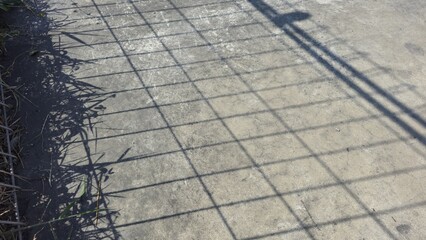 Concrete ground with the shadow of the fence. Rough sidewalk surface with shadows from metal fencing and a pole, creating a contrasting geometric pattern. 