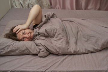 Caucasian mature female lying in bed looking distressed under blanket in bedroom setting.