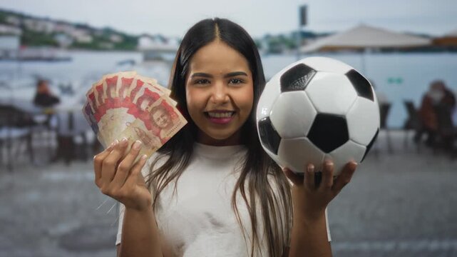 Young woman holding colombian pesos and soccer ball on outdoor restaurant terrace smiling happily with a scenic backdrop.
