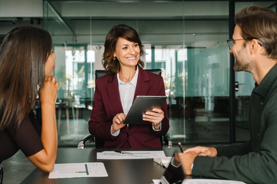 Smiling gesturing female corporate boss manager holding tablet, presents project to collaborative colleagues or client. Business mature CEO businesswoman leader leads diverse team meeting in office. 