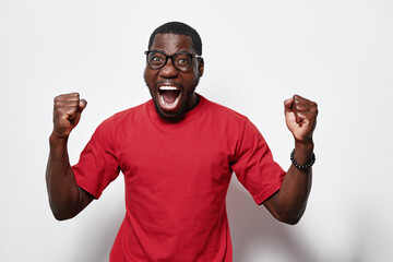 Fototapeta premium African American man with dark skin wearing glasses and red t shirt celebrating with fists up and mouth open on white background. Excited, happy, success and joy expression.