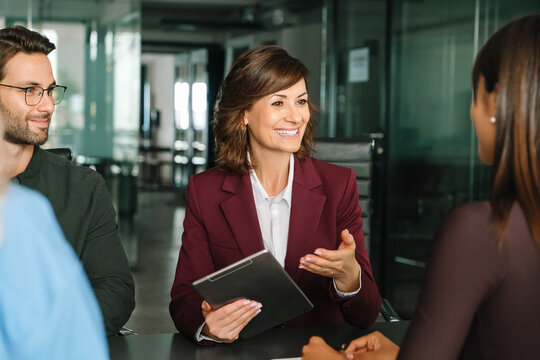Business mature CEO businesswoman leader leads diverse team meeting in office. Smiling gesturing female corporate boss manager holding tablet, presents ideas to collaborative colleagues or client 