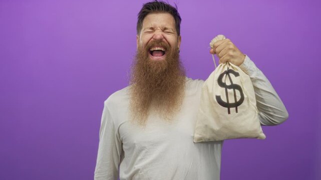 Bearded man smiling and pointing at canvas moneybag printed with dollar sign in purple studio; excitement.