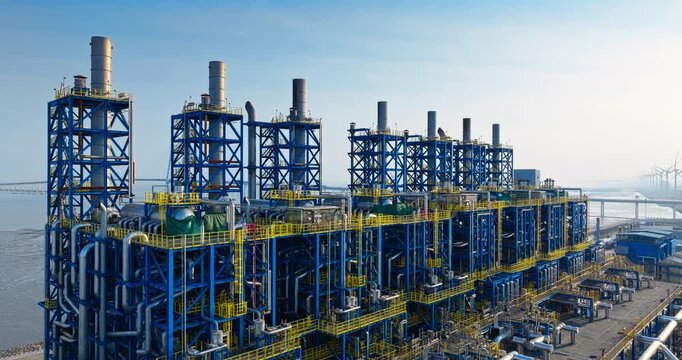 Large industrial chemical plant with blue steel structures and tall chimneys in a coastal area during the day.