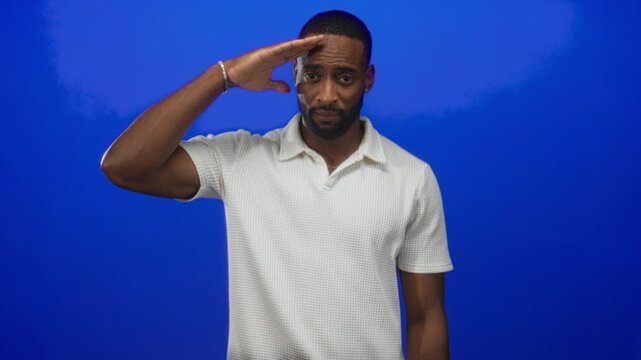 Man saluting with hand to forehead showing bare forearm and bracelet in studio against vibrant cobalt blue backdrop; respect duty.