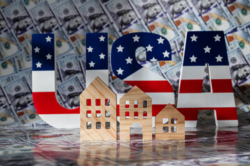 Obraz premium Symbolic wooden houses and the letters U S A with the American flag against the backdrop of $100 bills