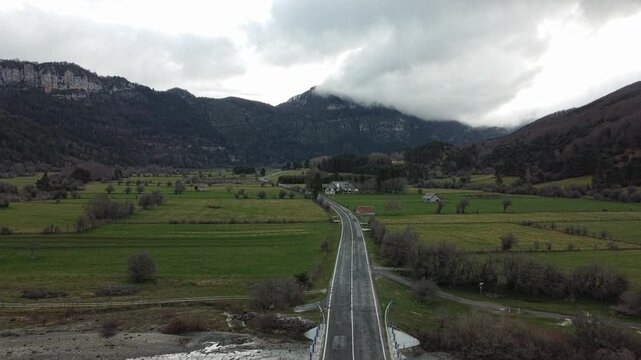 Rural landscape of the Roncal Valley, Isaba, Navarre, Spain, seen from an aerial view. A straight road crosses a green valley with meadows and open fields, surrounded by wooded hills and mountains. 