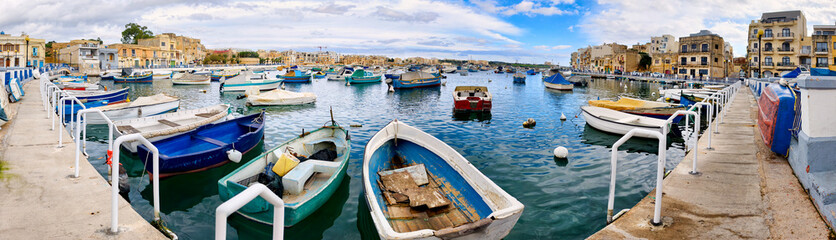 Panoramic view of Birzebbuga's small harbor with numerous moored boats and a lively promenade  rows of houses appear in the background. © EKH-Pictures