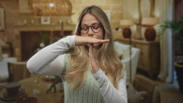 Blonde woman with glasses and long hair shows hands forming time out sign in hotel room; pause break reset hush.