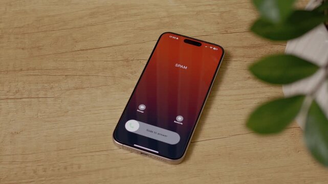 A smartphone receives an incoming call marked as spam. The phone is on a wooden table, showing the screen clearly with the spam label.