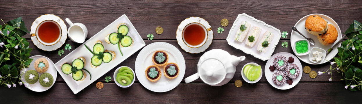 St Patrick's Day tea party table scene against a dark wood banner background. Variety of green shamrock theme finger sandwiches, fruits, scones and sweet desserts. Above view.