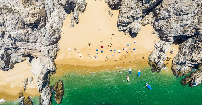 Top down aerial view of Playa de los Amantes in Los Cabos, Baja California Sur. Capturing the secluded beach framed by rocky cliffs, turquoise waters, anchored boats, and visitors enjoying the sand.