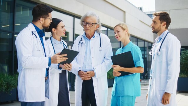 Cooperative medical team standing outside hospital. Caucasian male and female doctors discussing charts. Senior male physician observing colleagues. Nurse sharing updates near clinic building.