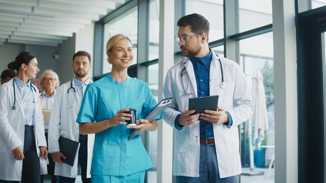 Focused nurse holding coffee and clipboard while walking with calm expression through clinic hallway. Male doctor beside her reviewing notes on tablet and preparing tasks. Team moving forward.