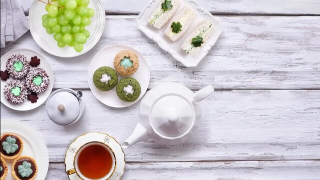 St Patrick's Day tea party table setting against a white wood background. Stop motion video. Variety of green shamrock theme finger sandwiches, fruits, scones and sweet desserts. Top down view.