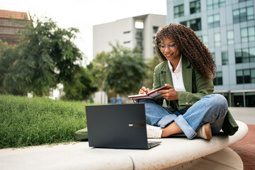 Young African American female student using laptop learning sitting in university garden park,...