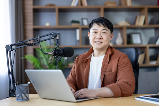 Asian man smiling, recording a podcast on his laptop with a professional microphone in a comfortable home studio, creating and broadcasting audio content