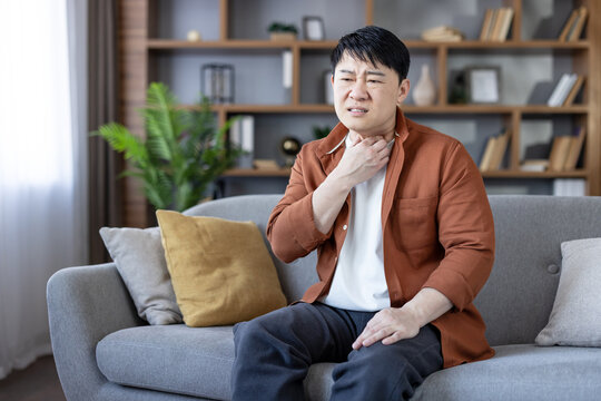 Asian man sitting indoors on a sofa, experiencing throat pain and discomfort while touching his neck, representing a common illness or allergic reaction symptom