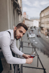 Naklejka premium Man holding a glass of red wine while leaning on a balcony in a city
