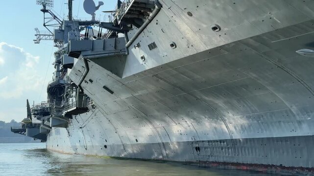 The radar mast and superstructure of the USS Intrepid rise above the deck in New York harbor. Antennas, satellite dishes, and steel structures show the ship's naval technology against a blue sky.