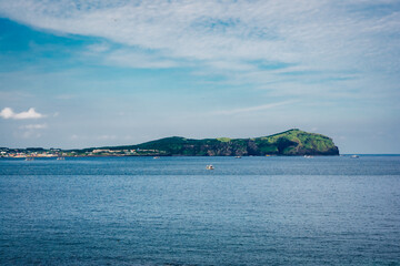 Naklejka premium A wide coastal seascape on Jeju Island, South Korea, with calm blue water and a green volcanic island on the horizon. Small boats dot the ocean under a bright sky with soft, textured clouds.