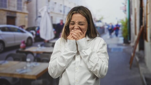Woman with hands covering mouth and fingers clasped, eyes closed, wearing white shirt on a restaurant terrace outdoors by sidewalk tables and umbrellas; surprise reaction.