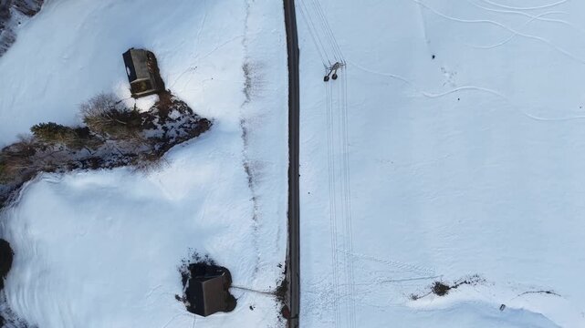 Top-down drone flight over a secondary mountain road with small curves, patchy snow on the edges, visible ski tracks on the snow, power lines crossing, and perpendicular alpine valleys, Swiss Alps win