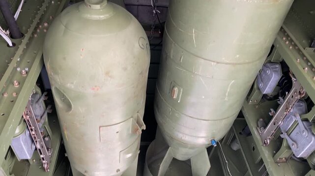 Inside view of a vintage bomber aircraft shows the bomb bay structure and metal framework. Cylinders, brackets, and panels reveal detailed military aviation engineering.