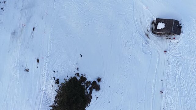 Top-down drone flight over a ski lift station where skiers line up for the drag lift, others descend on the slope beside it, icy and patchy snow visible on the piste, Swiss Alps winter resort.