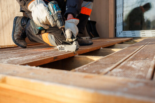 A worker in insulated gloves fastens grooved deck boards to timber joists with a cordless drill and hidden clips outside a modern house, frost on wood, warm side light.