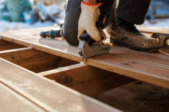 A gloved worker kneels on timber joists, fastening grooved cedar planks with a pneumatic stapler. Frost on boards and diffused golden light suggest cold outdoor work.