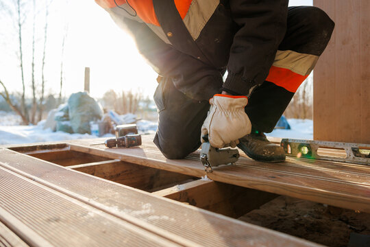 A worker in insulated gear kneels fastening composite boards to a timber frame using a pneumatic nailer, with a spirit level and cordless drill nearby in winter light.