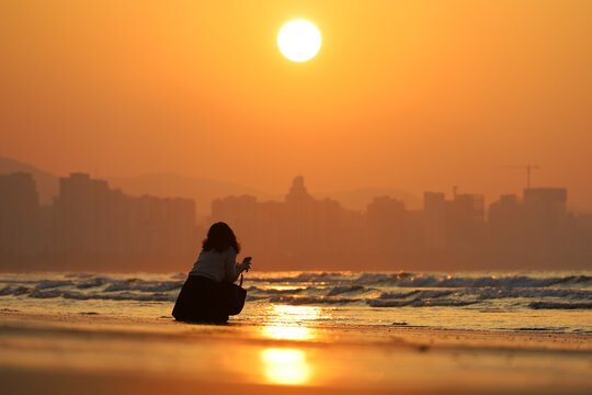 Silhouette of woman with smartphone on sea beach on city background. Sunrise on a coast, travel and holiday concept