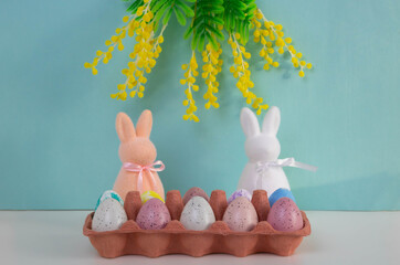 On a blue background, Easter eggs are placed in a container on a table. Pink and white rabbit toys are placed next to the mimosa branches. The concept of Easter.