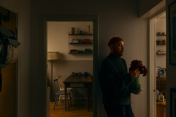 A calm bearded man holding a vase of flowers stands in a cozy, softly lit apartment interior. The scene captures a quiet moment of everyday life, warmth, and modern home living. © Francis