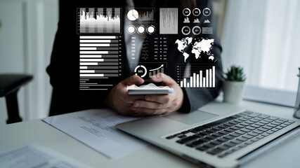 A business professional engages in data analysis using a smartphone while seated at a desk with a laptop, showcasing visual charts and statistics in a contemporary office setting. Impute