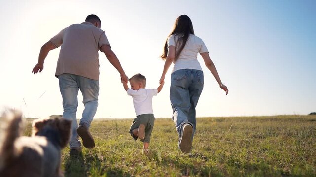 A man, woman, and boy run hand-in-hand through a tall grassy field. The boy skips as the man and woman hold his hands in the sunny grassy field under the blue sky.