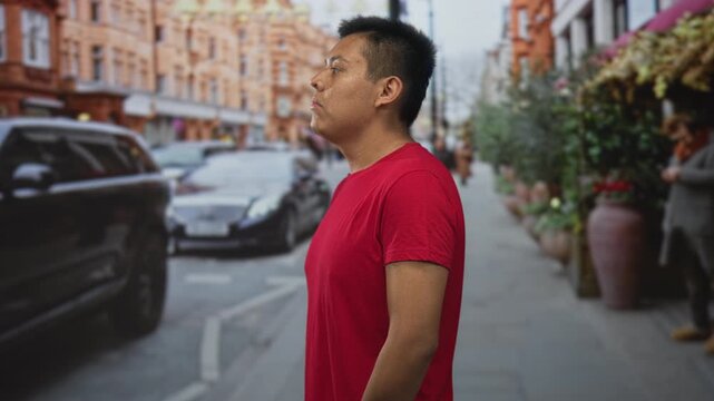 Man in red t shirt standing in profile on street with arms relaxed at sides, looking ahead toward traffic and shopfronts; urban solitude.