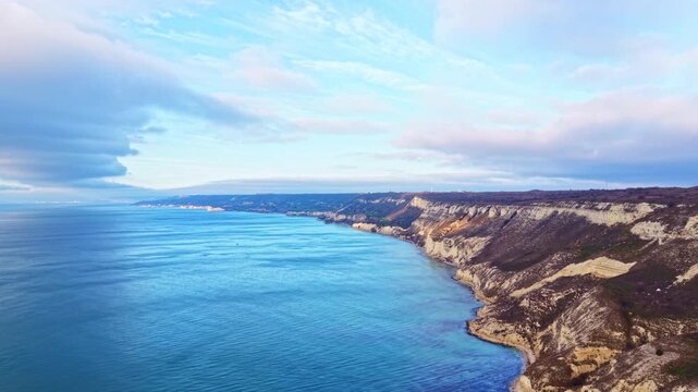 Water meets land where cliffs rise above the sea. Waves gently move along the shore under a cloudy sky. The scene captures a stretch of coastline without any buildings or people.