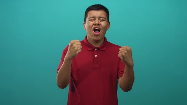 Young man with clenched fists and open mouth in studio against teal wall, wearing red polo shirt; triumph.