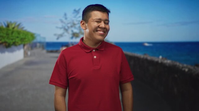 Man smiling in a red polo shirt on a coastal street promenade with hands relaxed at sides and shoulders square; calm contentment.