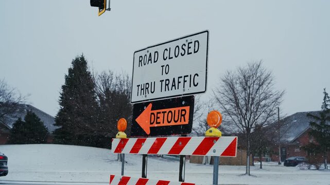 A road closed sign with detour notice stands on a snow-covered street. traffic control, road safety, transportation disruption caused by snowfall. Close POV view