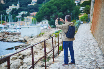 Mature tourist man with backpack photographing the scenic coastline of Villefranche-sur-Mer near Nice, France
