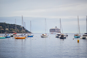 Fototapeta premium Scenic view of colorful sailboats and yachts anchored in calm blue sea with a large white cruise ship in the background under a cloudy sky